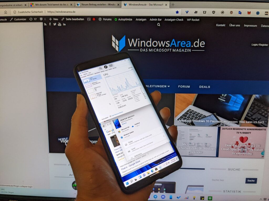 Foto eines Smartphones, auf dem Windows 11 läuft, direkt vor der WindowsArea-Webseite, die im Hintergrund auf einem Monitor geöffnet ist.